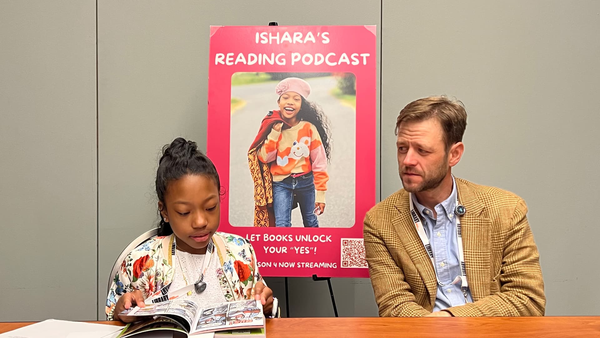 Ishara reading a graphic novel at the interview table beside Mac Barnett