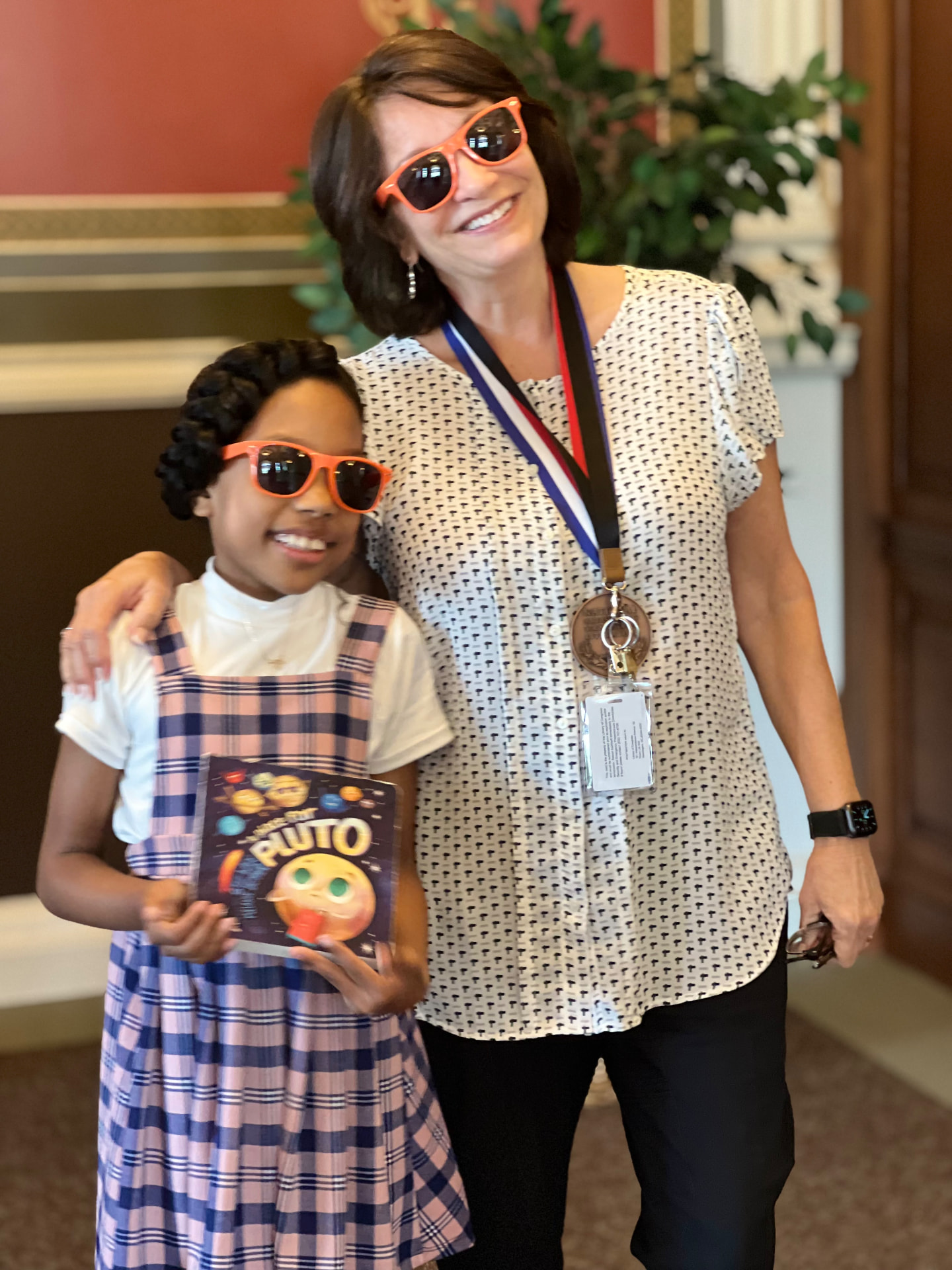 Ishara holding the book Pluto while posing with Meg Medina, both wearing orange sunglasses