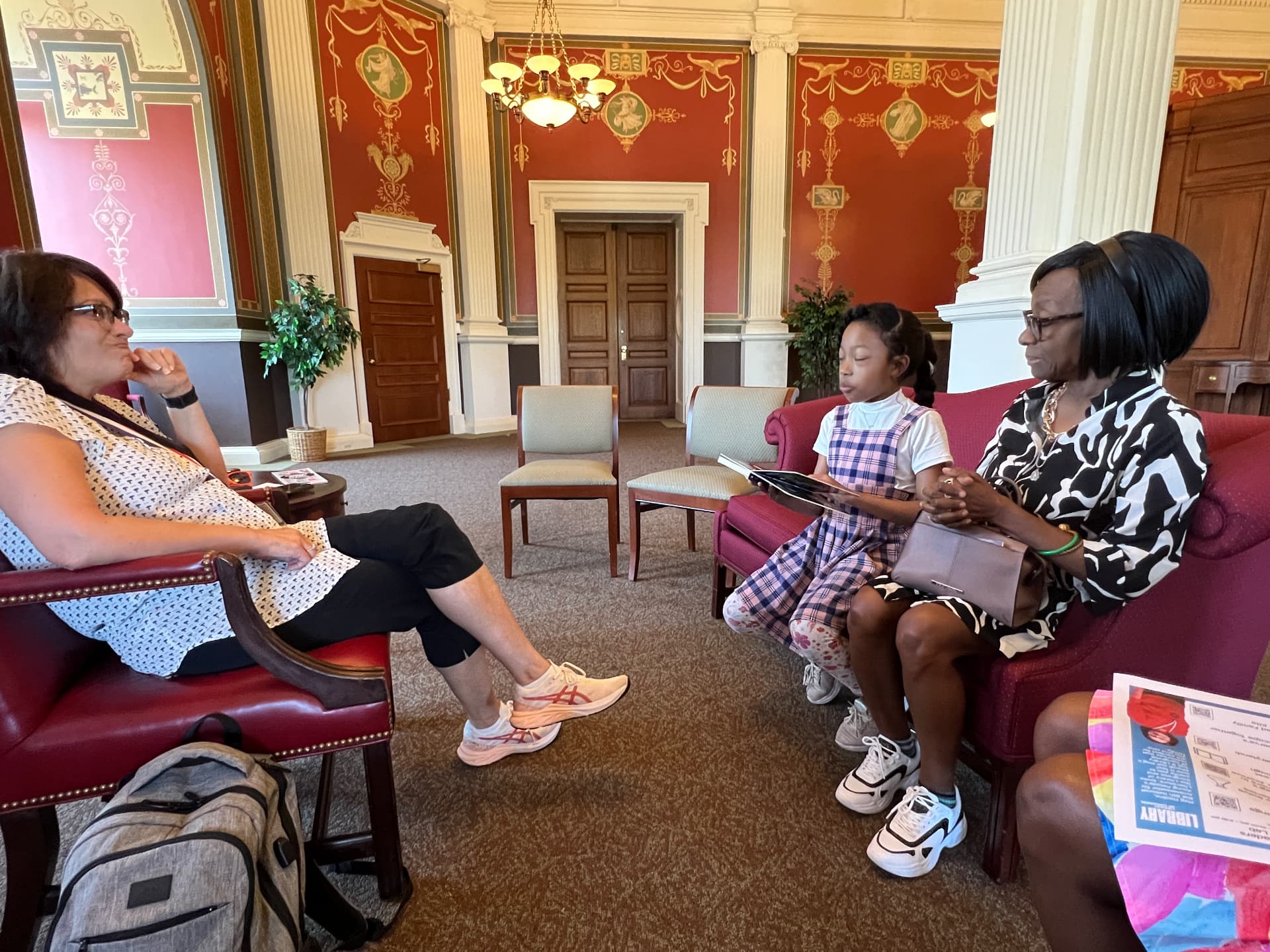 Ishara reading aloud from a book on a sofa at the Library of Congress with two adults listening