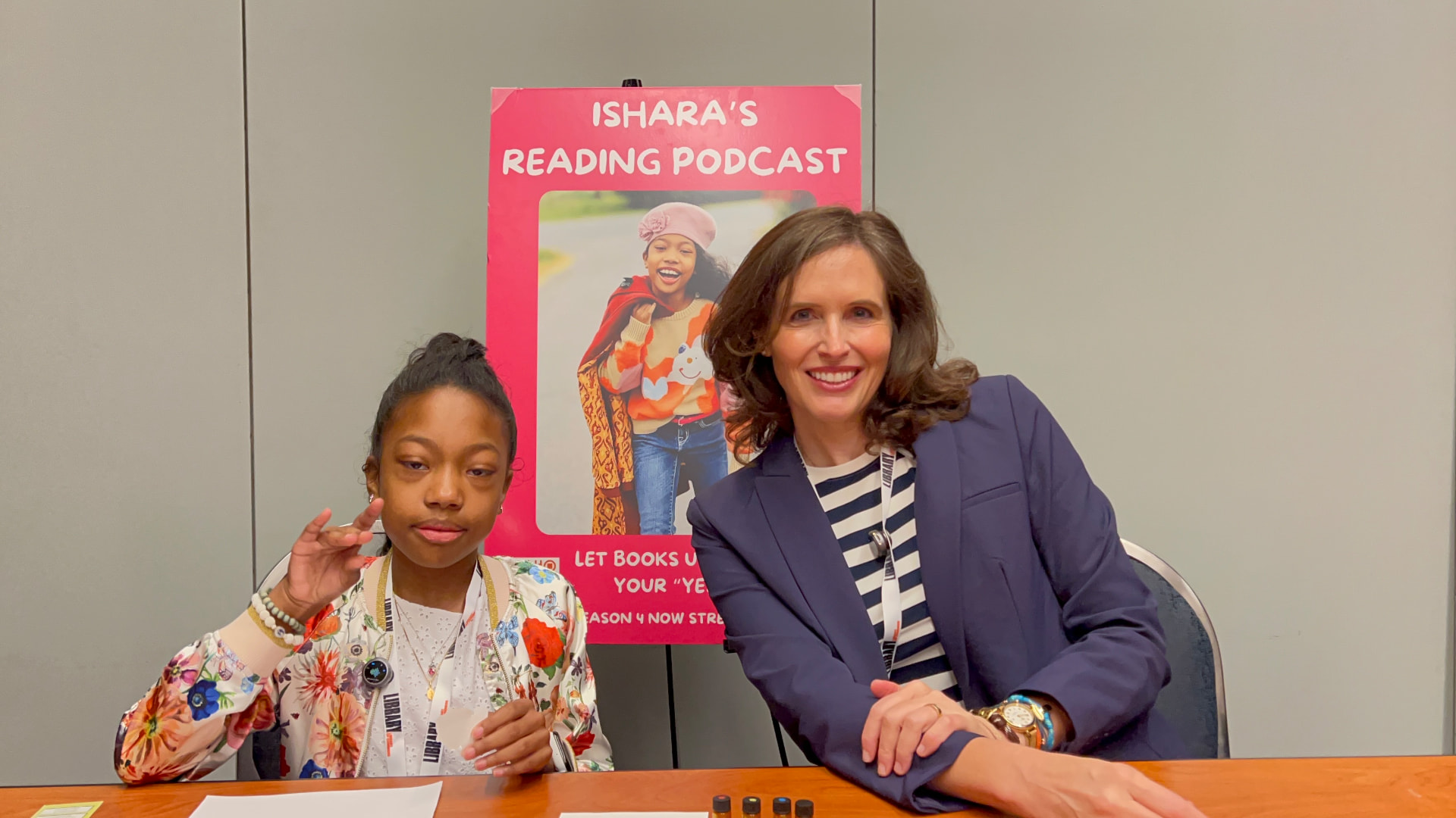 Ishara sitting at an interview table with Dr. Eleanor Spicer Rice in front of the Ishara's Reading Podcast poster